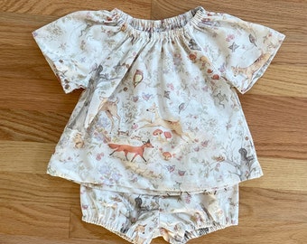 May include: A white cotton baby outfit with a short-sleeved top and bloomers. The top has a gathered neckline and a woodland animal print. The bloomers have an elastic waistband and a ruffled hem.