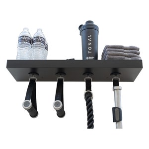 May include: A black wall-mounted fitness shelf with attached exercise handles, a rope, and a bar. The shelf holds two water bottles, a black shaker bottle with "TONAL" text, and a stack of gray towels. The handles have gray grips.