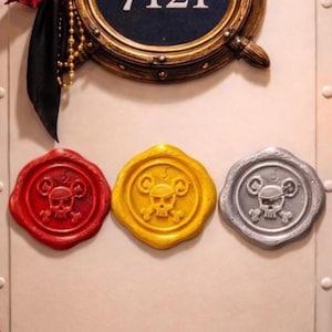 Op de afbeelding: Drie decoratieve waszegels in rood, goud en zilver, elk met een schedel- en gekruiste botten-ontwerp. De zegels zijn bevestigd op een licht oppervlak, met een rond bord erboven.