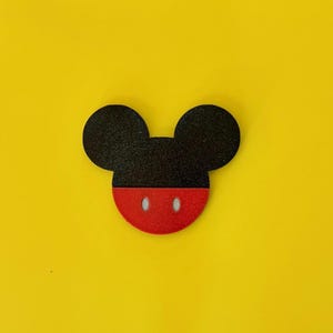 May include: A black and red Mickey Mouse-shaped design on a yellow background. The design features a black head with two round ears and a red face with two white oval-shaped eyes.