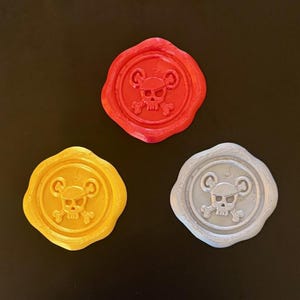 May include: Three wax seals in red, gold, and silver, each featuring a skull and crossbones design. The seals have a circular shape with a decorative border, and are set against a dark background.
