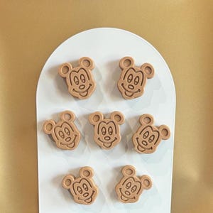 May include: Seven light brown Mickey Mouse-shaped magnets are arranged on a white, arched background. The magnets have a detailed face design and are made of a solid material. The background is a solid, light brown color.