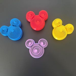 Könnte beinhalten: Vier farbenfrohe, 3D-gedruckte Mickey Mouse-Kopf-Formen in Blau, Rot, Gelb und Lila. Jede Form besteht aus konzentrischen Kreisen, die einen geschichteten Effekt erzeugen. Die Formen sind auf einer dunkelgrauen Oberfläche angeordnet.