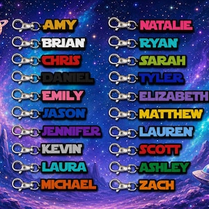 May include: A collection of twenty-two rectangular keychains with silver-toned lobster clasps, each featuring a unique name in bold, colorful lettering against a dark background, displayed over a vibrant purple and blue space-themed backdrop.