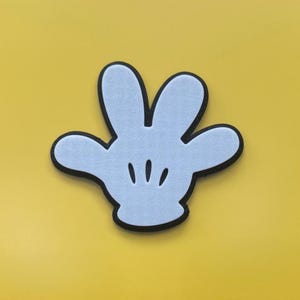 Puede incluir: Una mano de dibujos animados azul claro con un contorno negro y tres líneas cortas en la palma. La mano está sobre un fondo amarillo brillante. El diseño recuerda a un popular personaje de dibujos animados.