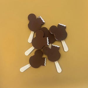 May include: A collection of chocolate-covered ice cream bars shaped like a famous mouse character. The ice cream is dark brown with a white bite mark, and each bar has a wooden stick. The background is a solid mustard yellow.