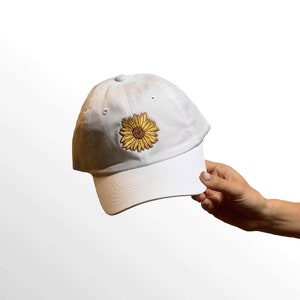 May include: White baseball cap with a yellow and brown sunflower embroidered on the front. The cap has a curved brim and a button on top. The cap is held by a person's hand.