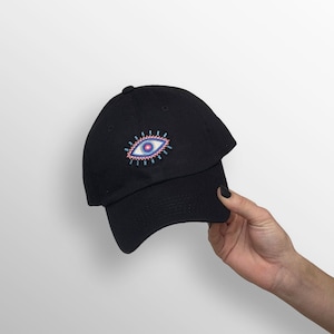 May include: Black baseball cap with an embroidered eye design in pink, blue, and white. The cap is made of a soft material and has a curved brim. The eye design is centered on the front of the cap.