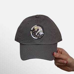 May include: A gray baseball cap with a circular patch featuring a black cat, a white cat, a crescent moon, and a sun design. The cap is made of a soft material and has a curved brim.