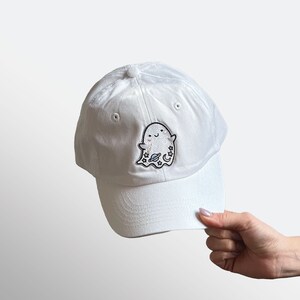 May include: White baseball cap with a curved brim. The cap features an embroidered patch of a smiling ghost with stars and a crescent moon. The cap is made of a soft, smooth fabric.