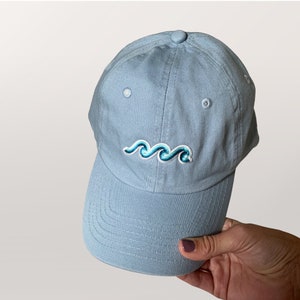 May include: Light blue baseball cap with a curved brim. The cap features an embroidered wave design in shades of blue. The cap is made of a soft fabric and has a button on top.