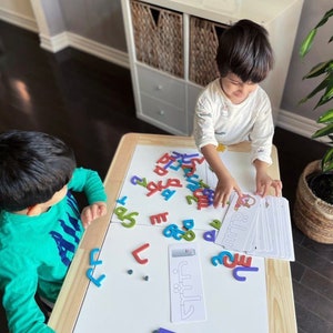 May include: Two children are sitting at a white table with a wooden frame. They are playing with colorful foam letters and cards with Arabic script. The letters are scattered on the table. The cards have pictures of animals and the Arabic word for the animal.