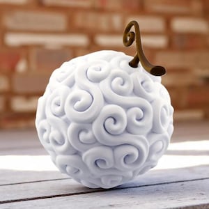 May include: A white, spherical object with a swirling, textured surface and a brown, curved stem. The object is set against a blurred background of a brick wall and wooden surface. This item is a decorative piece.