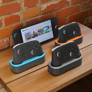May include: A black handheld gaming console with a screen displaying game graphics, along with three black game controllers in colored charging docks. The docks are in blue, orange, and gray. The controllers have white accents.