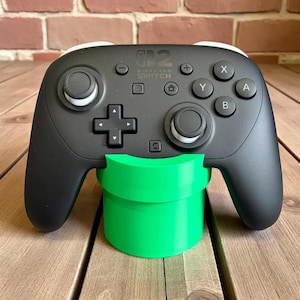 May include: A black Nintendo Switch Pro Controller resting on a bright green pipe-shaped stand. The controller has white accents and the standard button layout. The stand is made of plastic and is designed to hold the controller upright.