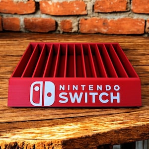 May include: A red Nintendo Switch game cartridge holder. The holder has multiple vertical slots and the Nintendo Switch logo in white. The holder is on a wooden surface with a brick background.