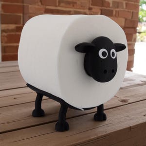 May include: A novelty toilet paper holder shaped like a sheep. The white roll of toilet paper forms the body, with a black sheep head and legs. The holder is designed to sit on a flat surface, adding a whimsical touch to any bathroom.