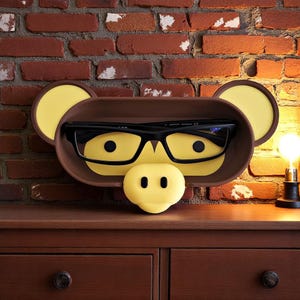 May include: A brown and yellow monkey-shaped eyeglass holder with black-framed glasses inside. The holder has yellow ears and a yellow nose with two black dots. The holder is on a wooden surface in front of a brick wall.