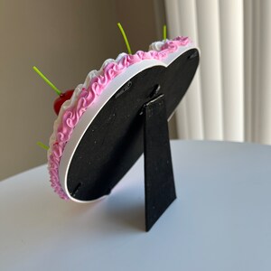 K&ouml;nnte beinhalten: Ein wei&szlig;er herzf&ouml;rmiger Bilderrahmen mit rosa Zuckerguss und einer roten Kirsche oben drauf. Der Rahmen hat einen schwarzen St&auml;nder.