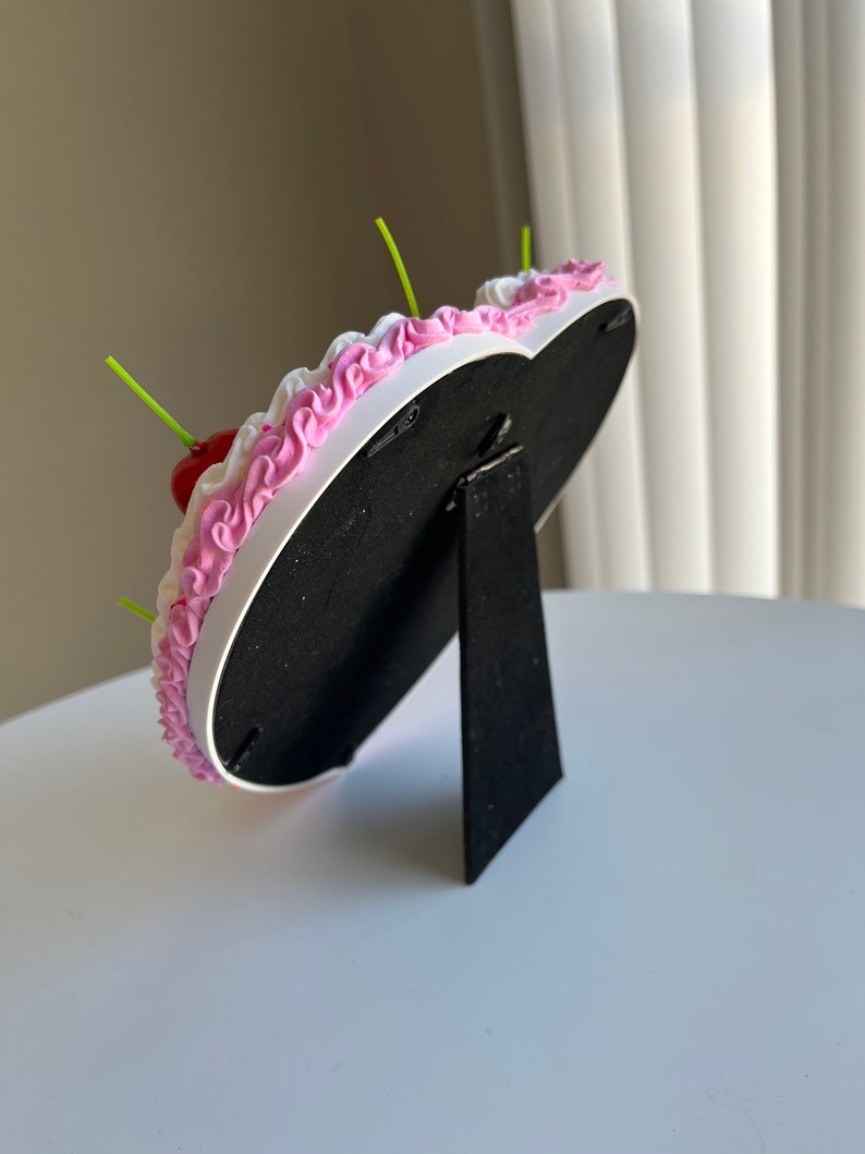 K&ouml;nnte beinhalten: Ein wei&szlig;er herzf&ouml;rmiger Bilderrahmen mit rosa Zuckerguss und einer roten Kirsche oben drauf. Der Rahmen hat einen schwarzen St&auml;nder.