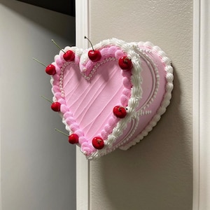 Könnte beinhalten: Ein rosa herzförmiger Kuchen, der mit weißem Zuckerguss und roten Kirschen verziert ist. Der Kuchen hängt an einer Wand.