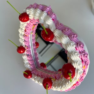 K&ouml;nnte beinhalten: Ein herzf&ouml;rmiger Spiegel mit einem rosa und wei&szlig;en Zuckerguss-Design und roten Kirschen am Rand.