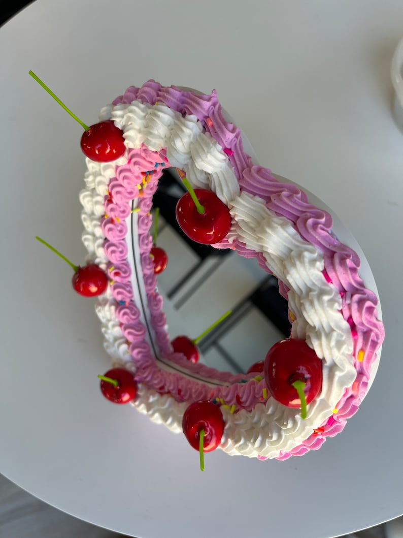 K&ouml;nnte beinhalten: Ein herzf&ouml;rmiger Spiegel mit einem rosa und wei&szlig;en Zuckerguss-Design und roten Kirschen am Rand.
