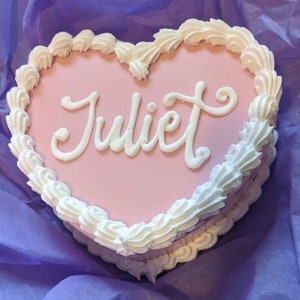 Może przedstawiać: Różowe ciasto w kształcie serca ozdobione białym lukrem i napisem "Juliet" napisanym białym lukrem.