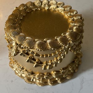 Puede incluir: Un pastel redondo de color dorado con glaseado texturizado y adornado. El pastel presenta una parte superior lisa y glaseado decorativo en los bordes y los lados, creando una apariencia lujosa. El pastel es de un solo nivel.