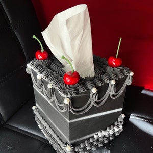 May include: Black and white tissue box cover shaped like a cake with white frosting and red cherries on top.