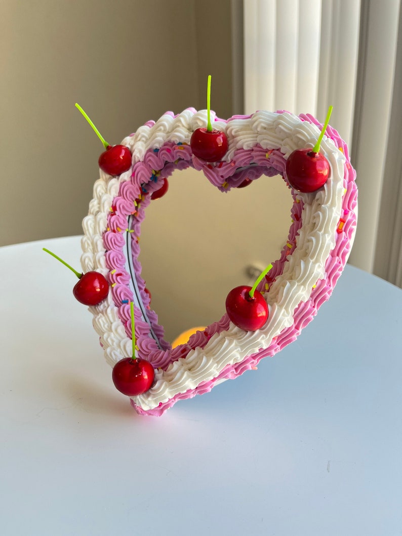 K&ouml;nnte beinhalten: Ein herzf&ouml;rmiger Spiegel mit rosa und wei&szlig;em Frosting-Design, verziert mit roten Kirschen und bunten Streuseln.