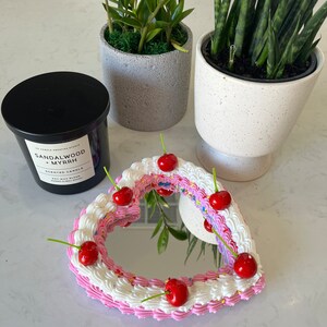 K&ouml;nnte beinhalten: Ein herzf&ouml;rmiger Spiegel mit einem rosa und wei&szlig;en Zuckerguss-Design, verziert mit roten Kirschen und bunten Streuseln. Der Spiegel ist von einer schwarzen Kerze mit dem Text "SANDALWOOD MYRRH SCENTED CANDLE SOY WAX BLEND HAND POURED IN CANADA" umgeben.