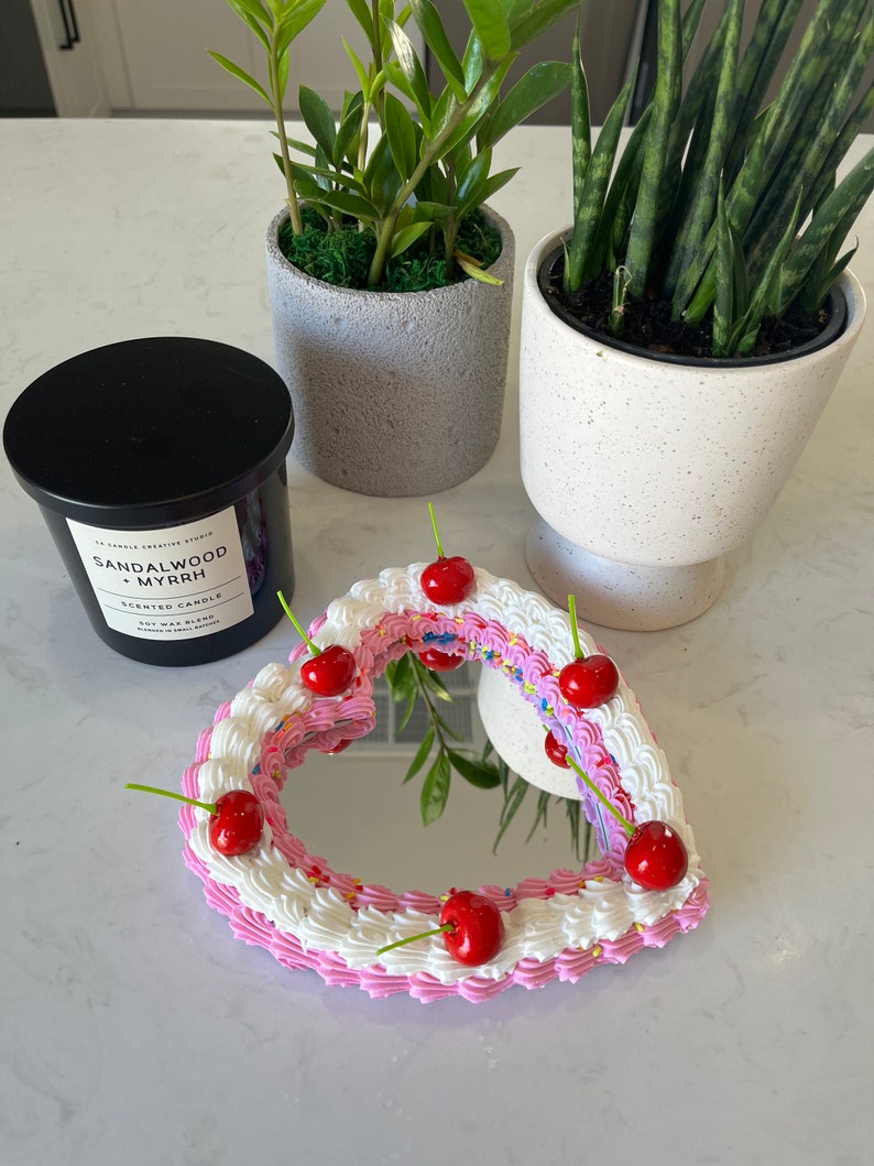 K&ouml;nnte beinhalten: Ein herzf&ouml;rmiger Spiegel mit einem rosa und wei&szlig;en Zuckerguss-Design, verziert mit roten Kirschen und bunten Streuseln. Der Spiegel ist von einer schwarzen Kerze mit dem Text "SANDALWOOD MYRRH SCENTED CANDLE SOY WAX BLEND HAND POURED IN CANADA" umgeben.