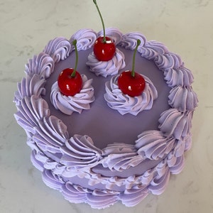 Puede incluir: Un pastel morado con glaseado blanco y tres cerezas rojas en la parte superior.