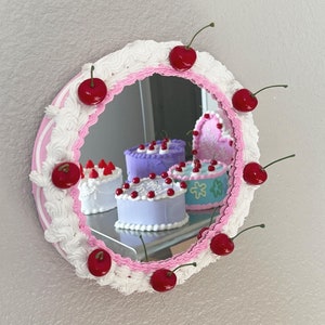 May include: A round mirror with a pink and white frosting design, surrounded by red cherries. The mirror reflects a variety of colourful cakes with frosting and cherries.