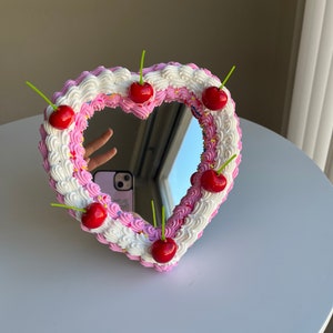 K&ouml;nnte beinhalten: Ein herzf&ouml;rmiger Spiegel mit einem rosa und wei&szlig;en Zuckerguss-Design und roten Kirschen am Rand.