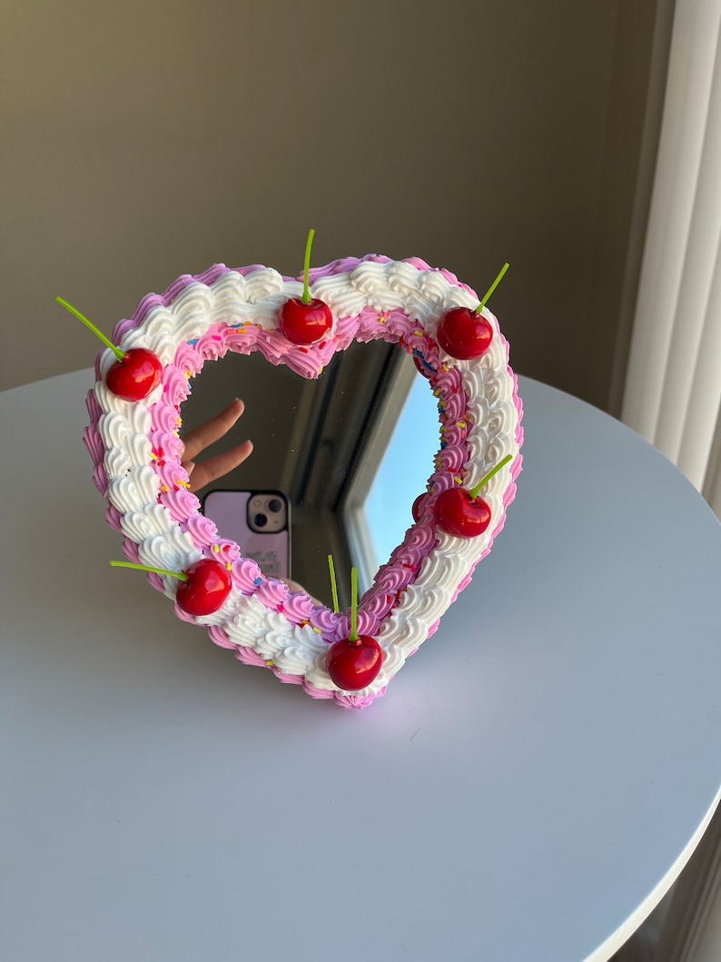 K&ouml;nnte beinhalten: Ein herzf&ouml;rmiger Spiegel mit einem rosa und wei&szlig;en Zuckerguss-Design und roten Kirschen am Rand.