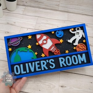 May include: A blue wooden sign with a space theme. The sign features a rocket, astronaut, planets, and stars. The text "OLIVER'S ROOM" is written in white letters on a blue background.