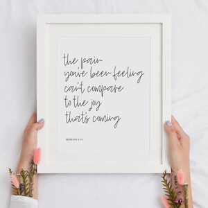 May include: White framed print with the text "the pain you've been feeling can't compare to the joy that's coming" and the scripture reference "ROMANS 8:18".