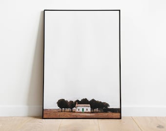 May include: Framed art print featuring a small white house with a green door, surrounded by trees. The artwork has a minimalist style, with a neutral color palette of white, brown, and green. The print is displayed on a wooden floor.