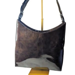 May include: A glossy, dark brown shoulder bag with a curved top and a single strap. The bag has a patent leather appearance and is displayed on a yellow stand against a white background. The strap is dark brown and has a buckle.