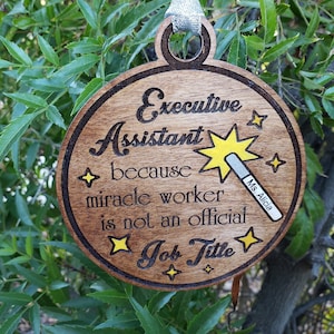 May include: A round wooden ornament with the text "Executive Assistant because miracle worker is not an official Job Title" and a yellow star with a wand. The ornament has a small name tag with the text "Ms. Alicia".
