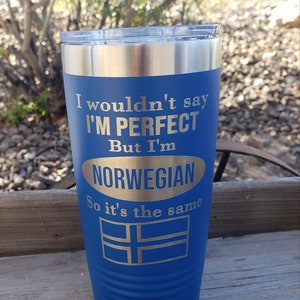 Könnte beinhalten: Ein blauer Edelstahlbecher mit einer weißen Aufschrift, die lautet "Ich würde nicht sagen, dass ich perfekt bin, aber ich bin Norweger, also ist es dasselbe". Der Becher hat einen silbernen Deckel und einen silbernen Band um die Oberseite. Die Aufschrift ist von einem silbernen Oval umgeben. Eine silberne norwegische Flagge befindet sich am Boden des Bechers.