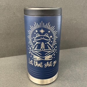 Puede incluir: Vaso aislado azul marino con tapa negra y base plateada. El vaso presenta un diseño grabado con láser de una rana meditando dentro de una estrella, con el texto "let that shit go".