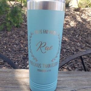 May include: A light blue insulated tumbler with a silver lid. The tumbler features the name "Rae" in a gold script font, surrounded by a floral wreath, and the text "SHE IS FAR MORE PRECIOUS THAN RUBIES PROVERBS 3:15".