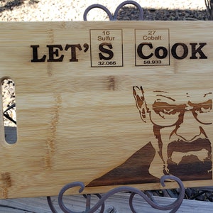 Peut inclure: Une planche à découper en bois avec un motif gravé d'un visage d'homme et le texte "LET'S COOK" avec la lettre "S" remplacée par le symbole chimique du soufre et le mot "COOK" remplacé par le symbole chimique du cobalt.
