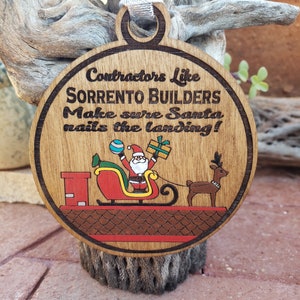 May include: Wooden Christmas ornament with a festive design. The ornament features Santa Claus in his sleigh, pulled by a reindeer, with the text "Contractors Like Sorrento Builders Make sure Santa nails the landing!" A silver ribbon is attached for hanging.