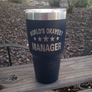 May include: A dark blue stainless steel tumbler with a silver lid. The tumbler has the text "WORLD'S OKAYEST MANAGER" with three stars printed on it.