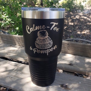 May include: Black stainless steel tumbler with a white design that says "Calme-Toe le Pompon". The design includes a white pom-pom hat.