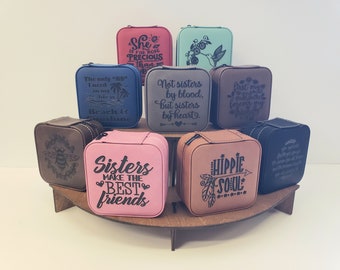 May include: Assortment of eight small square boxes with zipper closures. Each box is made of faux leather and features a different engraved design. The designs include a bee, a hummingbird, a palm tree, a heart, a feather, and various text phrases. The boxes are displayed on a wooden stand.